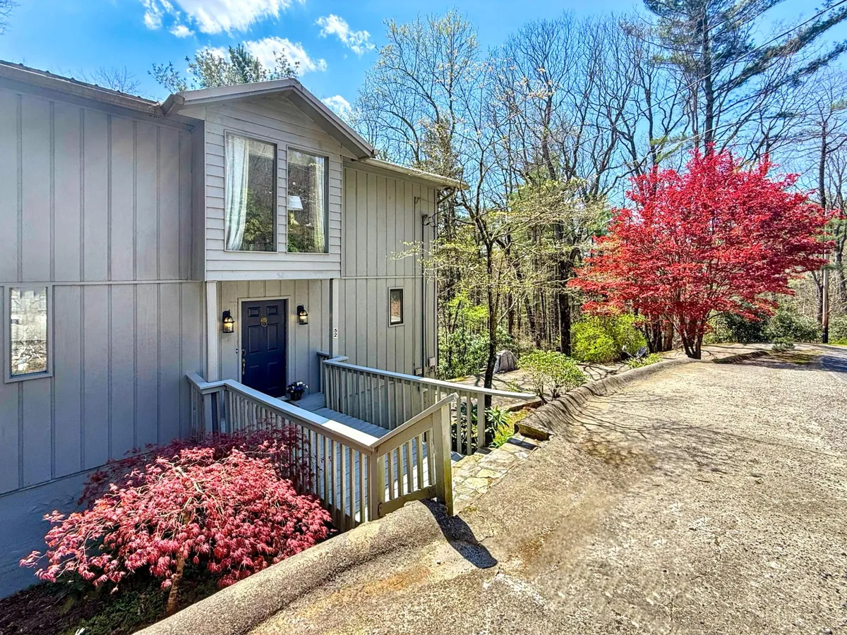The hero image showcases a charming single-family home nestled in lush greenery, with a striking blue front door, vibrant red flowering bushes flanking the entrance, and inviting wooden steps leading up to a welcoming porch. The scene captures the tranquil beauty of the Highlands, North Carolina setting, while the porch hints at leisurely mornings and peaceful afternoons spent outdoors.