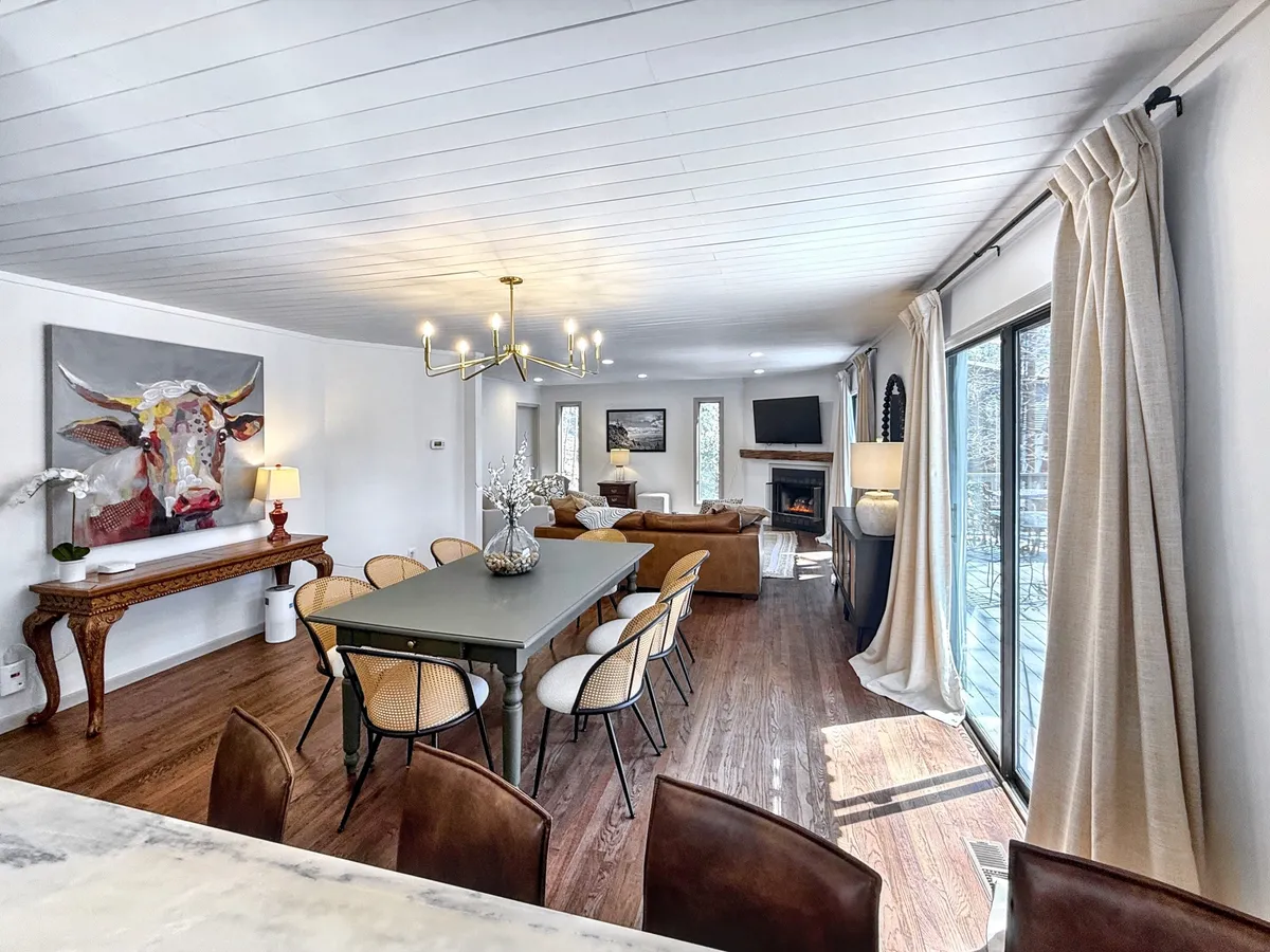 A welcoming dining area with a wooden dining table, elegant chandelier, playful cow painting, a cozy sofa area, and a gas log fireplace in the background. The setting is arranged for family dinners and intimate gatherings, with soft lighting enhancing the warmth.