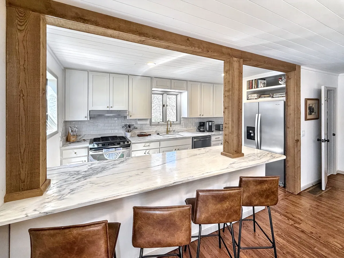 A close-up view of the kitchen area, featuring a marble countertop, wooden beam, modern kitchen appliances, and a row of bar stools by the breakfast bar. The scene is warm and inviting, perfect for gatherings and culinary creativity.