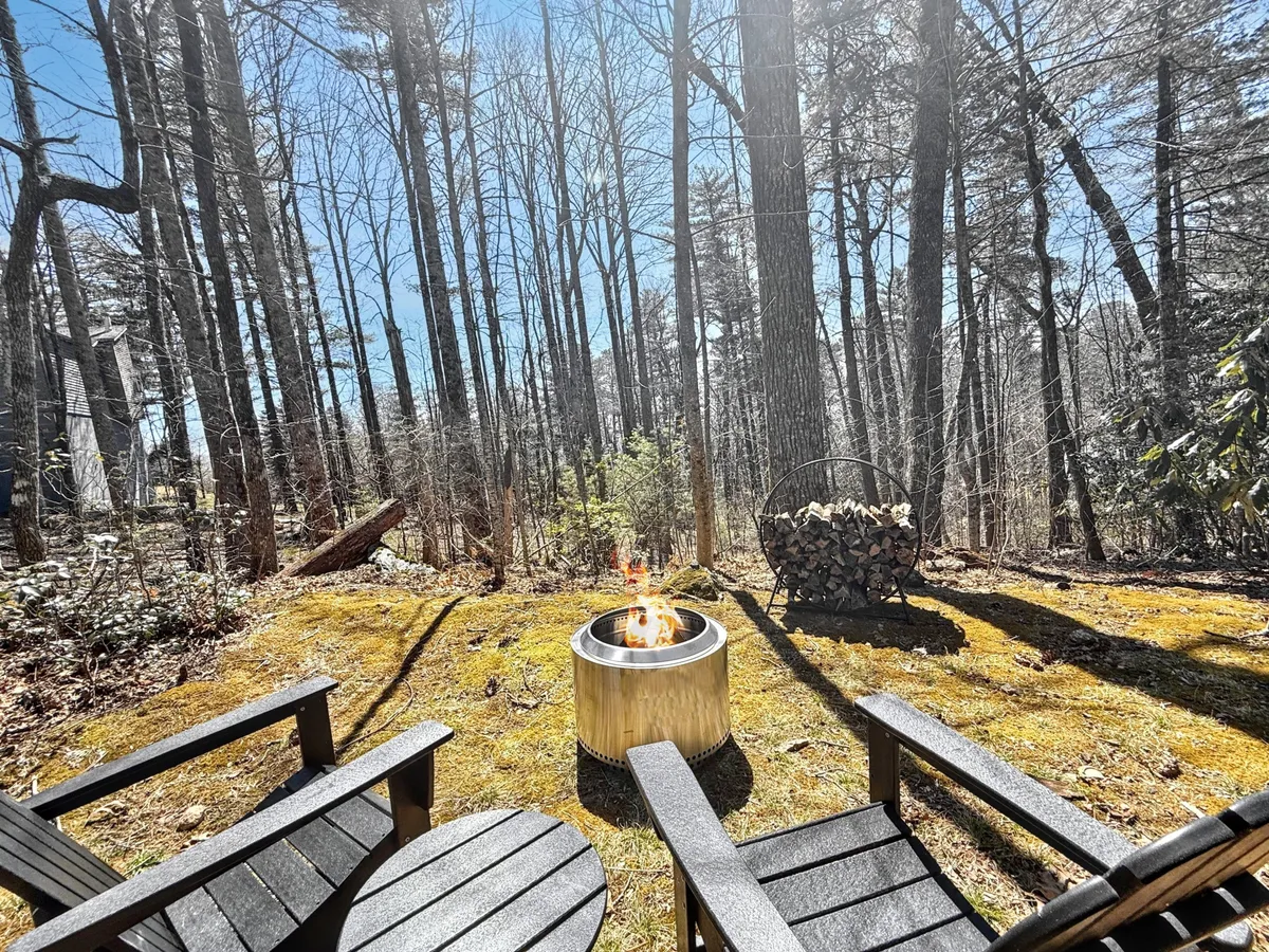 A cozy outdoor gathering spot featuring a fire pit surrounded by Adirondack chairs and a wood pile, all set amid tall trees. The setting suggests evenings spent relaxing, storytelling, or stargazing under the night sky. The environment combines comfort and rustic charm, blending relaxation with the outdoor experience.