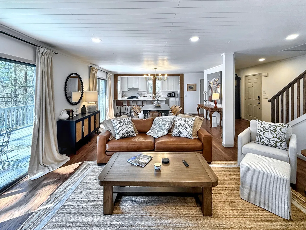 A bright, inviting living space featuring a comfortable sofa and coffee table set beside expansive windows. The windows frame lush mountain scenery, bringing the tranquil outdoors into the cozy interior. The adjacent dining area is perfect for transitioning between work and relaxation.