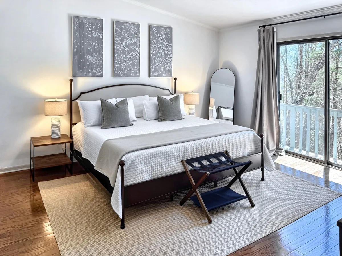A serene bedroom setting showcasing a comfortable bed, tasteful wall art, a sliding glass door leading to a private balcony, a nightstand, and a mirror. The soft tones and natural light create an inviting atmosphere, and the balcony access underscores the private sanctuary theme.