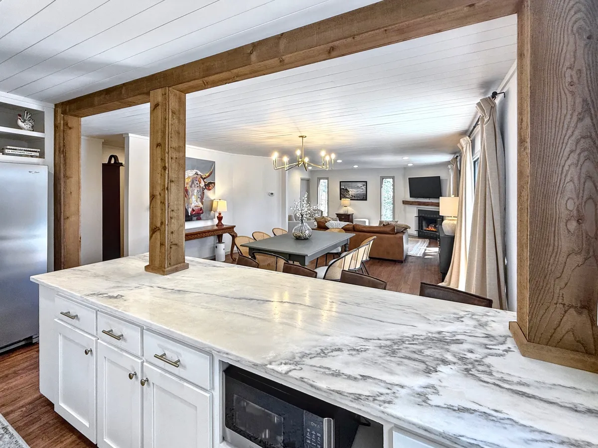 This image showcases the open-plan main living area, with a marble kitchen countertop, wooden beams overhead, a dining table set for entertaining, and the living area with a cozy sofa visible in the background. The layout is airy and flows seamlessly from one space to another.