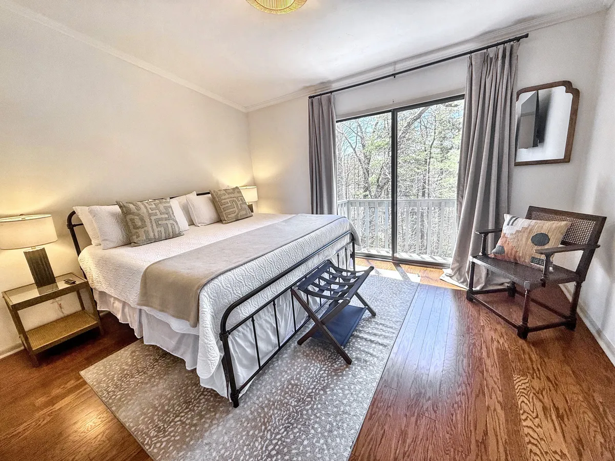 A tranquil king bedroom featuring a plush bed adorned with decorative pillows, large windows that flood the space with natural light, a stylish armchair for reading or relaxing, and a nightstand with a lamp. The view from the windows hints at the wooded, serene landscape beyond, and the overall décor is calming and sophisticated.